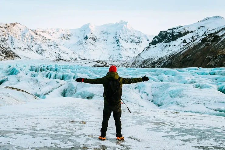 A traveler with outstretched arms stands on Langjokull Glacier in Iceland, surrounded by vast, icy formations and snow-capped mountains in the distance.