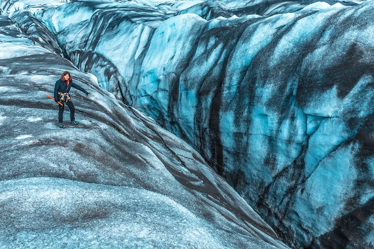 A hiker navigating the rugged and icy terrain of Svinafellsjökull Glacier in Iceland, showcasing the stunning blue and black ice formations.