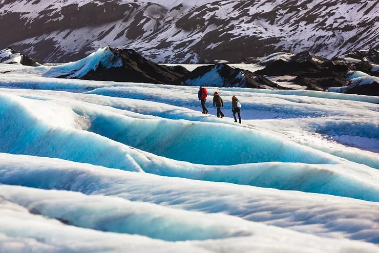 Three hikers exploring the stunning blue ice formations of Vatnajökull Glacier in Iceland, with rugged mountains in the background.