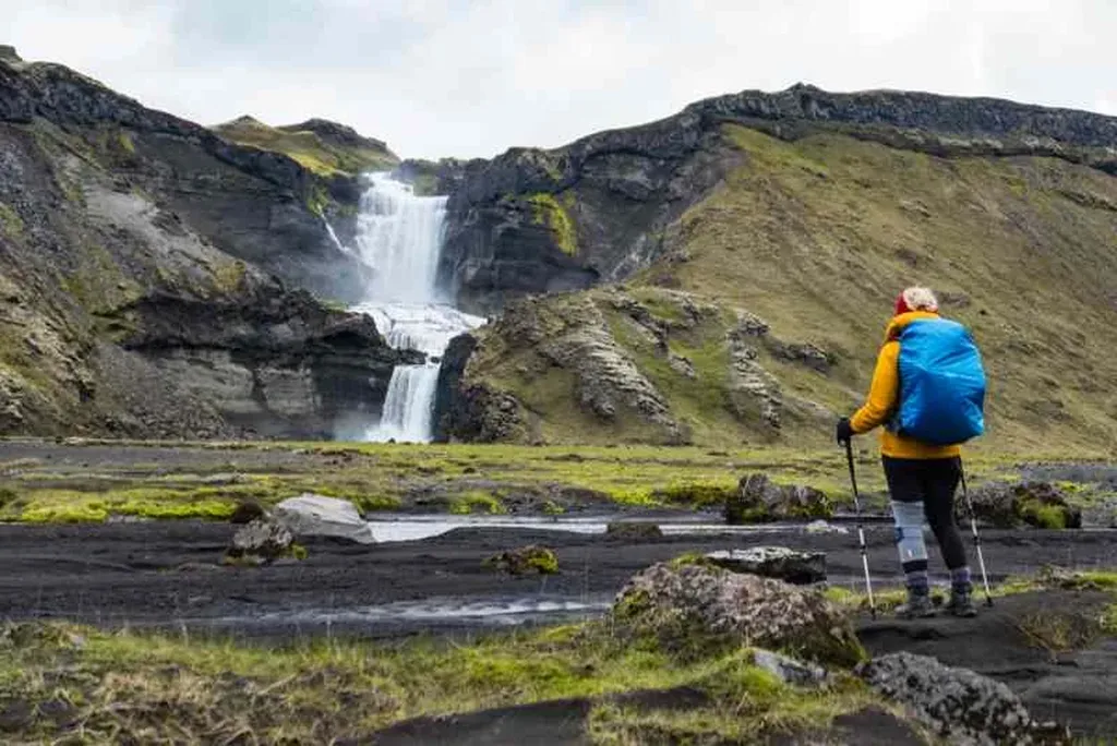 A hiker with a blue backpack and trekking poles standing in front of a majestic waterfall in Eldgjá, Iceland, capturing the spirit of adventure and the stunning natural beauty of the landscape.