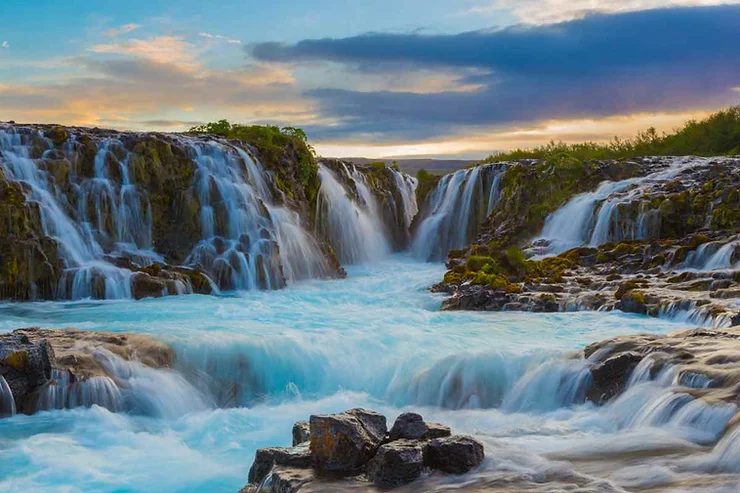 Bruarfoss Waterfall in Iceland, featuring cascading waters over jagged rocks into a vibrant, crystal-blue pool, set against the backdrop of a serene sunset sky with soft, colorful clouds.