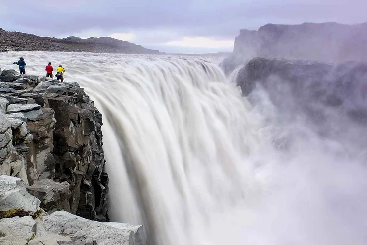 Three people in colorful rain jackets stand on a rocky cliff, gazing at the immense and powerful Dettifoss Waterfall, with water thundering down and mist rising into the air.