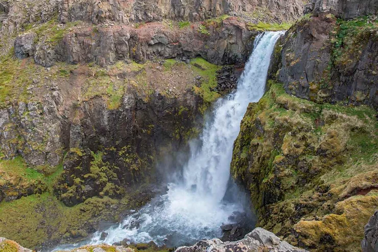 Gljúfursárfoss Waterfall nestled within rocky cliffs, with water cascading into a serene pool surrounded by lush green moss and rugged terrain.