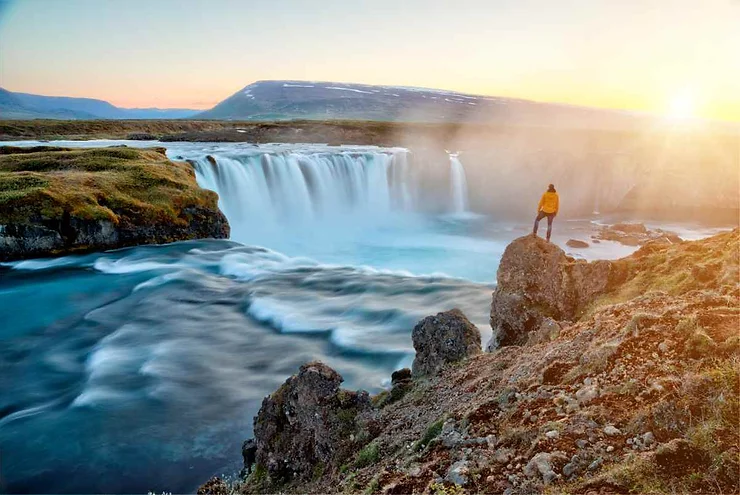 A person in a yellow jacket stands on a rocky outcrop, gazing at the majestic Godafoss Waterfall as it cascades into a serene river, with the sun setting in the background and casting a warm glow over the landscape.