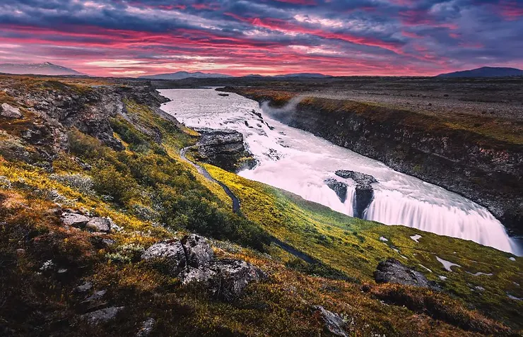 The majestic Gullfoss Waterfall in Iceland cascades powerfully into a rugged canyon, illuminated by a stunning sunset sky that paints the landscape in vibrant hues of pink and purple.