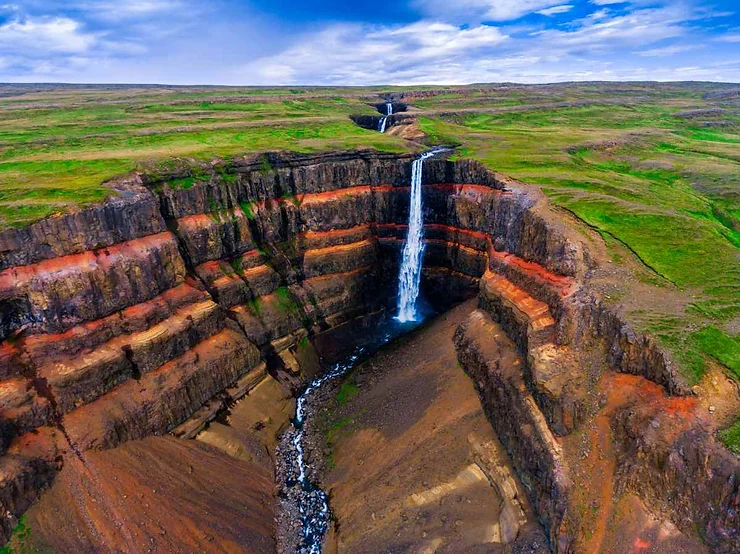 A breathtaking aerial view of Hengifoss waterfall cascading dramatically into a deep gorge, surrounded by layered rock formations and lush green plains under a bright blue sky with scattered clouds.