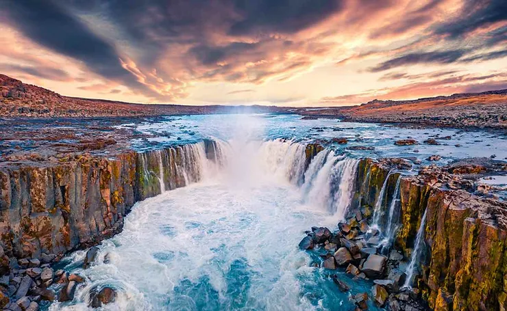 The breathtaking Selfoss Waterfall cascades over a rocky cliff into a circular basin, surrounded by rugged terrain, with a dramatic sunset sky painting the horizon.