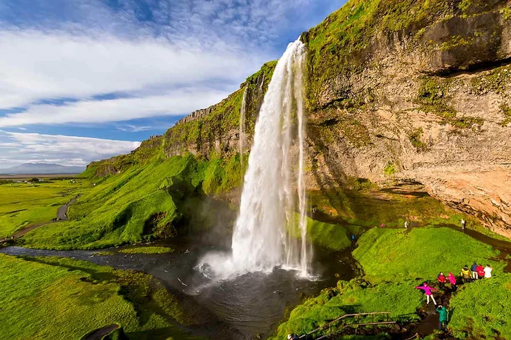 Seljalandsfoss Waterfall in Iceland, a stunning natural wonder with a powerful stream of water falling from a high cliff into a serene pool below, surrounded by lush green landscapes and clear blue skies, with visitors marveling at its beauty.