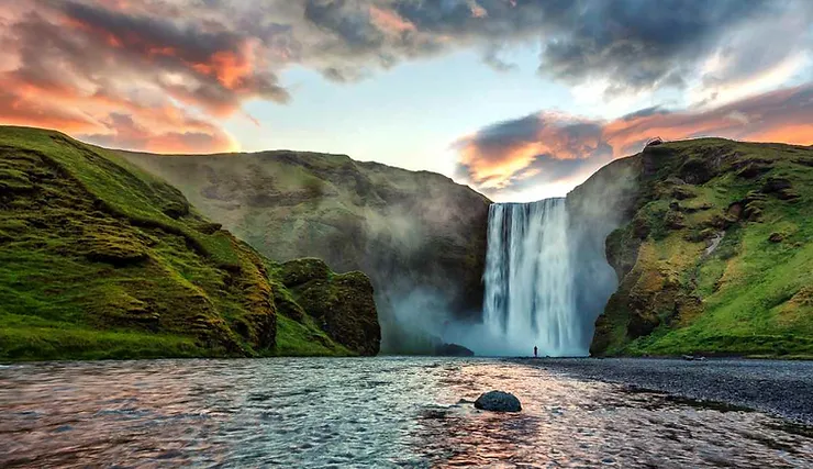 Skogafoss Waterfall in Iceland, a breathtaking sight with water powerfully cascading down a high cliff into a tranquil river, flanked by lush green hills under a dramatic sky painted with hues of orange and pink at sunset.