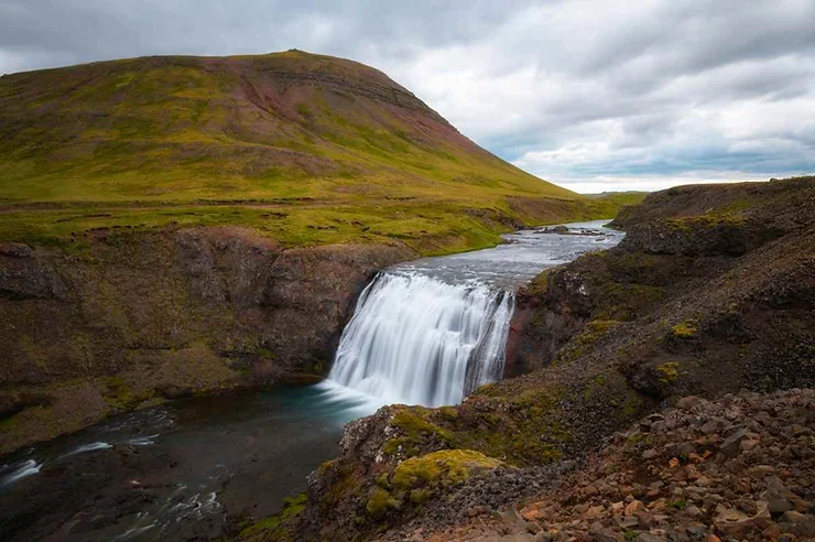 Thorufoss Waterfall in Iceland, showcasing a stunning cascade of water flowing over a rugged cliff into a serene river, surrounded by lush green hills under a cloudy sky.