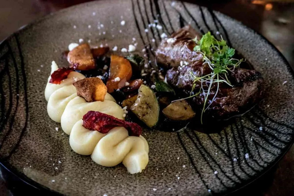 A beautifully plated dish of Icelandic cuisine is presented on a dark, textured plate. The dish features a portion of tender meat, garnished with fresh herbs, accompanied by a creamy mashed potato swirl and a variety of roasted vegetables. The vibrant colors and elegant presentation highlight the culinary artistry.