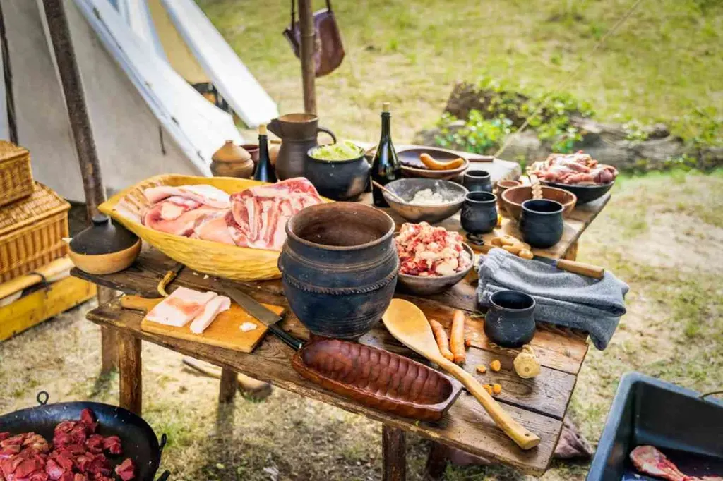 A rustic outdoor setup with various traditional Icelandic food items displayed on a wooden table. The table is laden with raw meats, bowls of ingredients, wooden utensils, and clay pots, evoking a historical or Viking-era feast.