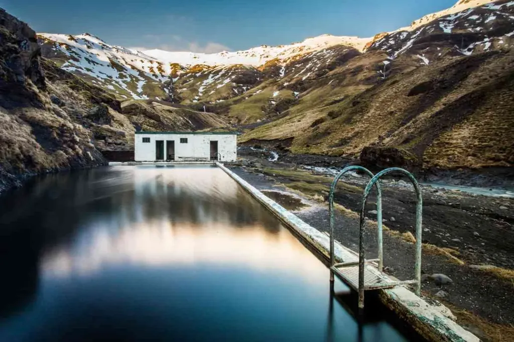 A hot pool in Iceland set against a stunning mountainous backdrop in February. The serene pool reflects the surrounding snow-capped mountains and the rustic white building at the edge of the pool, providing a tranquil and picturesque scene.