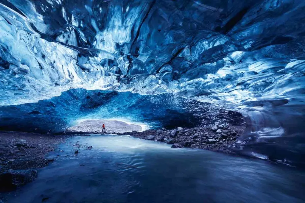 A person exploring an ice cave in Iceland during February. The cave's ceiling is a mesmerizing shade of blue with intricate ice formations, and a small stream runs through the cave, adding to the ethereal beauty of the scene.