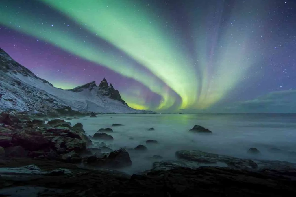 Stunning display of the Northern Lights over a snowy coastal landscape in Iceland during February, with vibrant green and purple auroras lighting up the night sky.