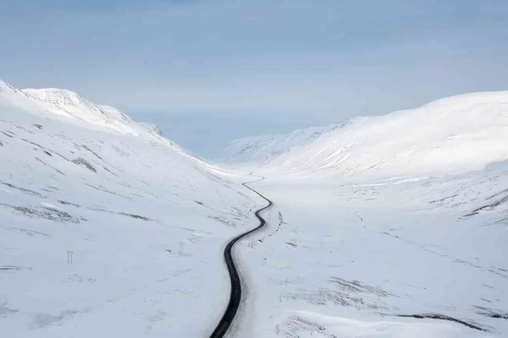 A winding road stretches through a vast, snowy valley in Iceland. The road is flanked by snow-covered mountains, creating a serene winter landscape. The sky above is clear with a hint of blue, adding to the peaceful atmosphere of the scene.