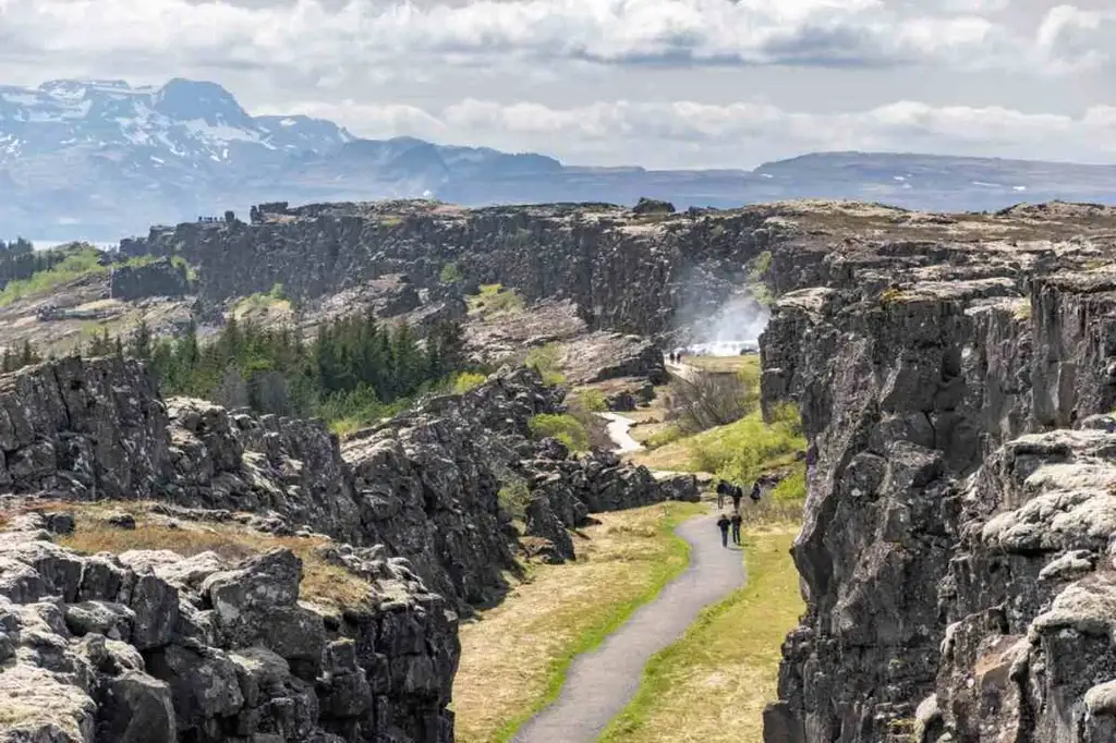 A scenic view of Almannagjá Gorge in Iceland, featuring a narrow pathway winding through rugged cliffs. In the background, there are snow-capped mountains under a partly cloudy sky. The gorge is surrounded by patches of green vegetation and trees, with a few people walking along the path.