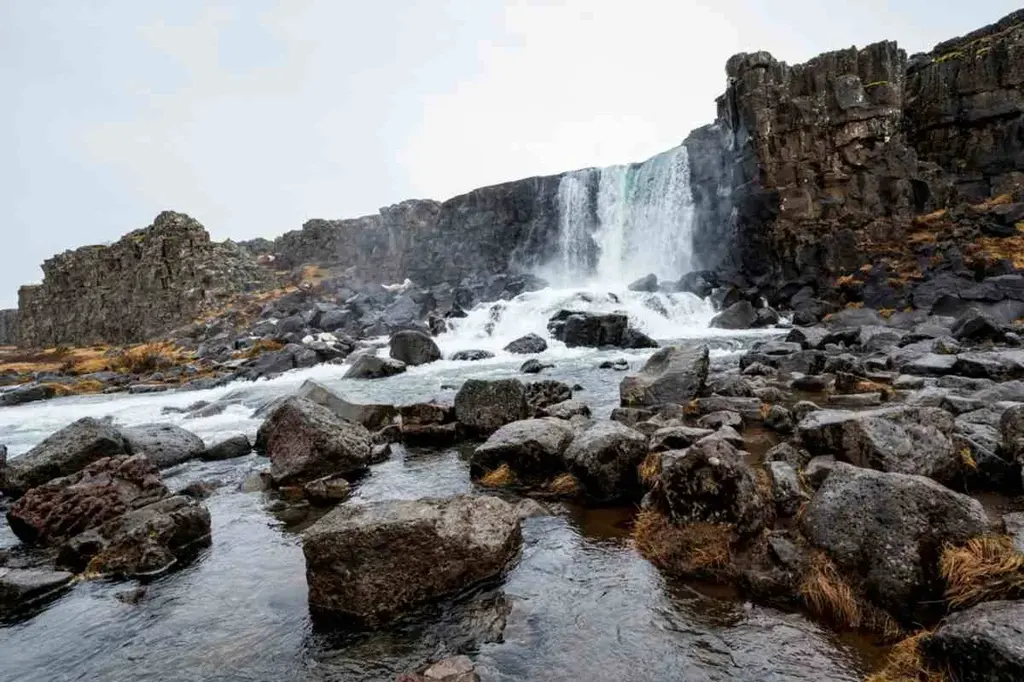 A stunning view of Öxarárfoss waterfall in Iceland, with water cascading down a rocky cliff into a pool surrounded by large boulders. The scene is set against a backdrop of rugged terrain, with mist from the waterfall creating a serene atmosphere.