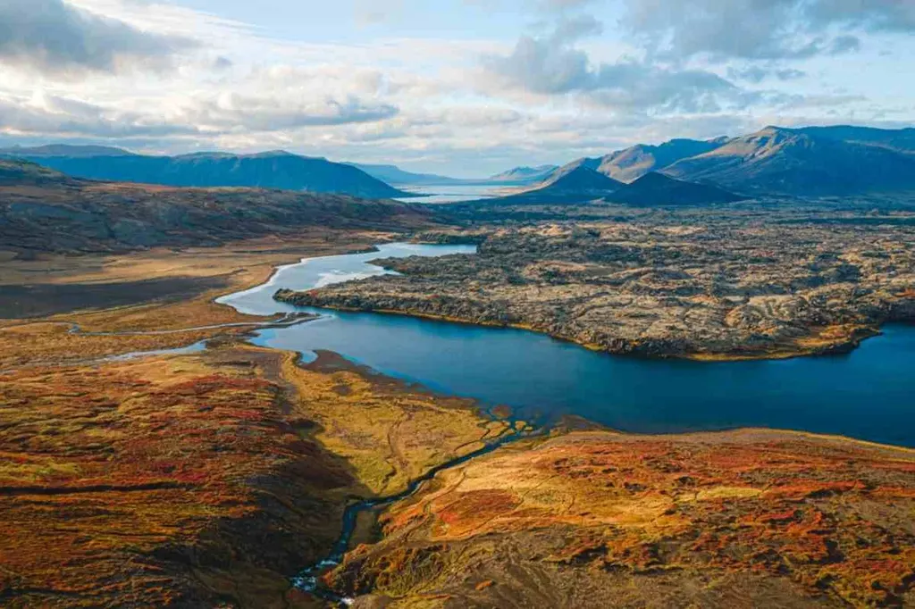 A breathtaking aerial view of Þingvellir National Park in Iceland, showcasing the vibrant autumnal colors of the landscape. The image captures the winding river and lake surrounded by rugged terrain, with distant mountains under a partly cloudy sky.