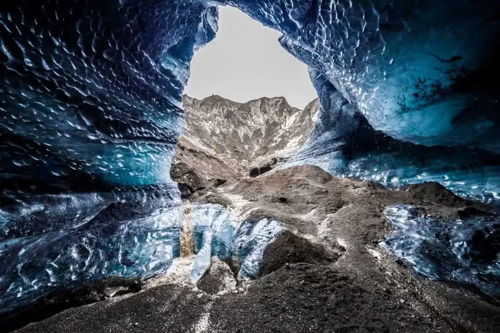 A stunning view from inside the Katla Ice Cave in Iceland, featuring vibrant blue ice walls that contrast sharply with the dark volcanic soil. The cave opens up to reveal a rugged mountain landscape under an overcast sky, highlighting the unique and dramatic beauty of this natural wonder.