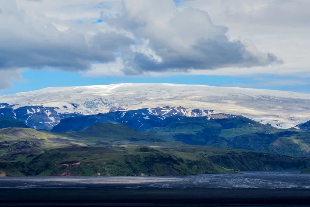 A picturesque view of the Katla volcano in Iceland, showcasing its snow-capped peak surrounded by lush green valleys and rugged terrain. The image is taken under a partly cloudy sky, with the vast landscape highlighting the natural beauty and dramatic contrasts of the Icelandic scenery.