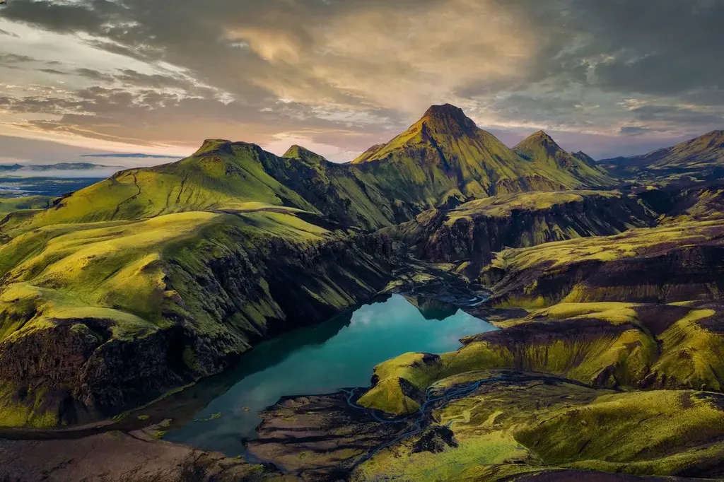 An aerial view of a stunning Icelandic landscape featuring lush green mountains and a serene blue lake nestled within a dramatic valley. The scene is bathed in the soft light of either dawn or dusk, highlighting the rich colors and textures of the terrain.