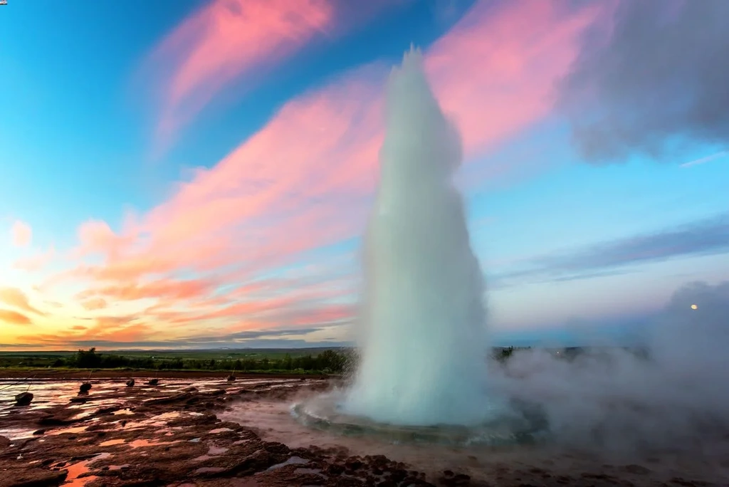 A powerful geyser eruption in Iceland, with water shooting high into the air against a vibrant sunset sky. The sky is painted in hues of pink, orange, and blue, creating a dramatic backdrop. The ground around the geyser is wet and rocky, reflecting the colors of the sky and the eruption.