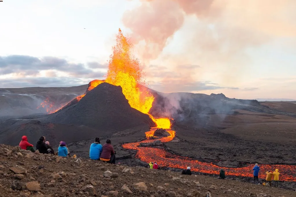 A group of people in colorful jackets sit and stand on rocky terrain, observing an active volcano erupting with bright, glowing lava flowing down its side. The scene captures the dramatic and powerful eruption, with plumes of smoke and ash rising into the sky, set against a backdrop of a serene Icelandic landscape under a softly lit sky.