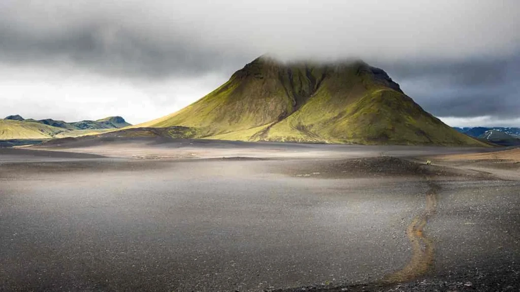 A dramatic view of a mountain in Thorsmork, Iceland, partially shrouded in low-hanging clouds. The mountain's slopes are covered in lush green moss, contrasting with the barren, dark volcanic plains that stretch out in the foreground. A faint trail winds through the desolate landscape, leading towards the imposing mountain.