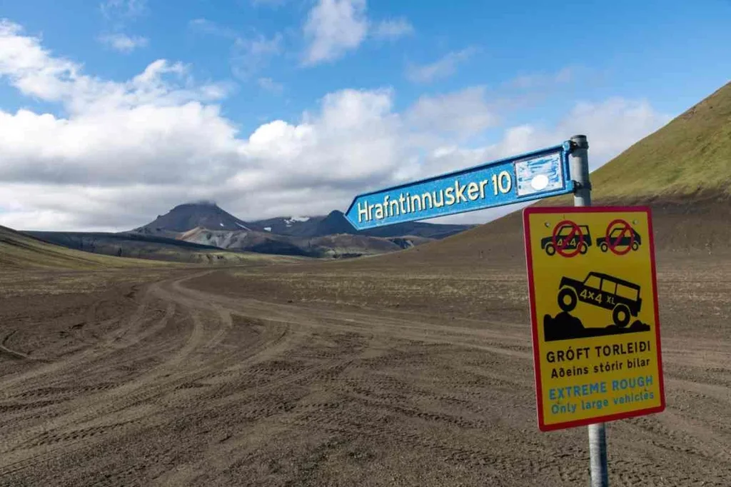 A dirt road in Thorsmork, Iceland, leading towards a distant mountain range under a bright blue sky with scattered clouds. A blue road sign indicates the direction to Hrafntinnusker while a yellow warning sign below alerts drivers about extreme rough terrain advising that only large 4x4 vehicles should proceed.