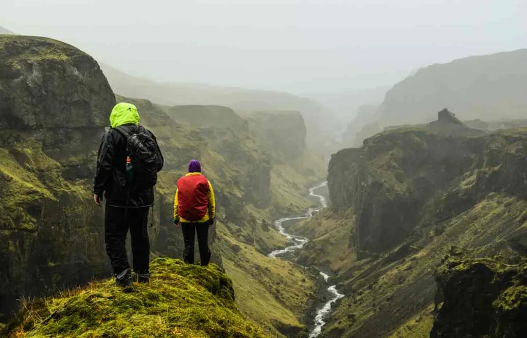 Two hikers stand on a grassy cliff edge in Thorsmork, Iceland, overlooking a dramatic canyon with steep, moss-covered cliffs and a winding river at the bottom. Both hikers are dressed in waterproof jackets, one in green and black and the other in red and yellow, as they take in the misty, overcast scenery.