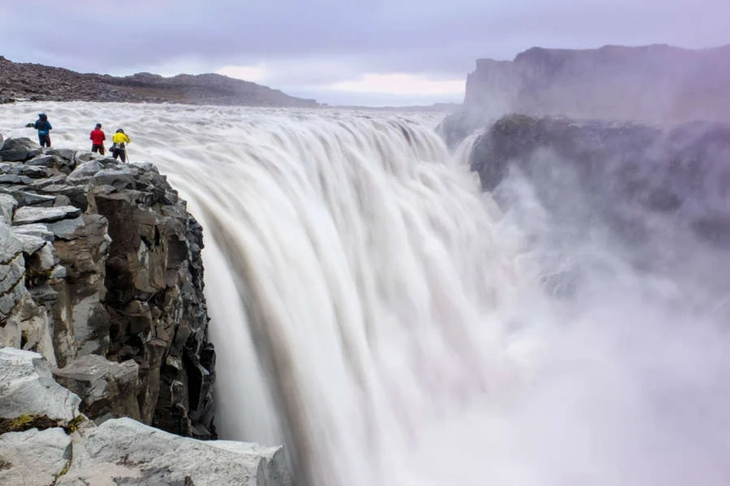 Three people in colorful jackets stand on the edge of a rocky cliff, looking at the powerful Dettifoss waterfall in Iceland. The massive waterfall plunges into the misty canyon below, creating a dramatic and awe-inspiring scene. The sky is overcast, adding a moody atmosphere to the landscape.