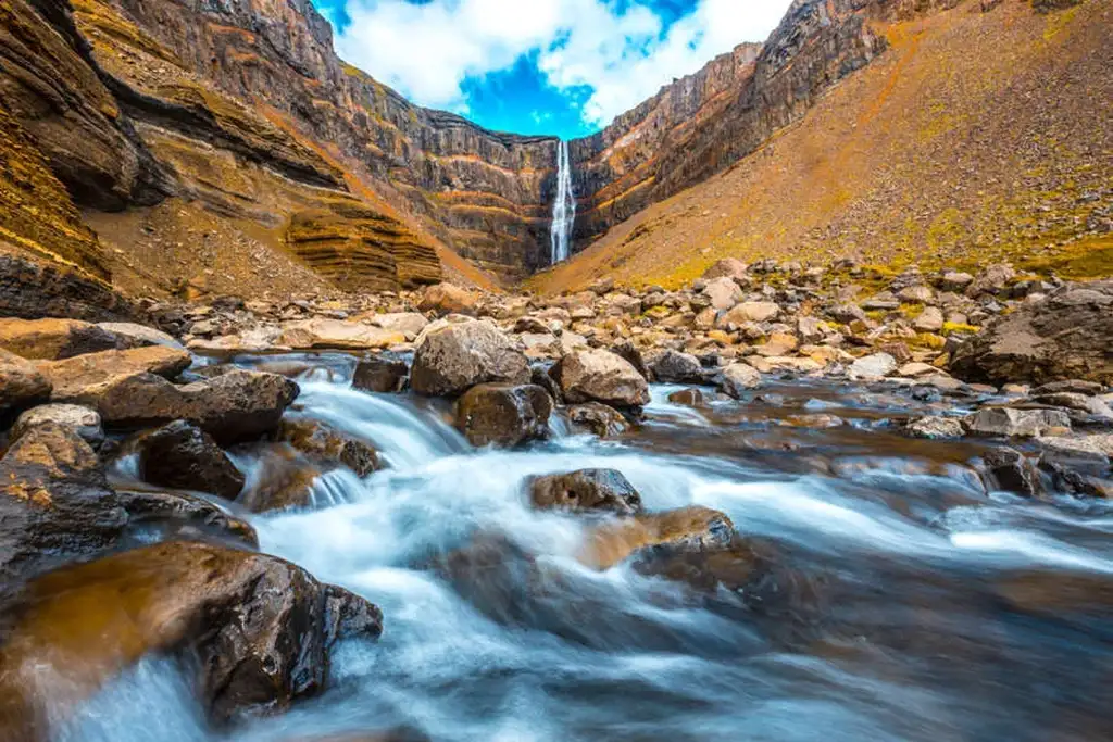 A dynamic view of Hengifoss waterfall in Iceland, with cascading water flowing down a tall cliff into a rocky stream. The scene is framed by rugged, steep canyon walls that showcase the geological layers in shades of brown, orange, and gray. The sky above is bright blue with scattered clouds, adding to the vibrancy of the landscape.