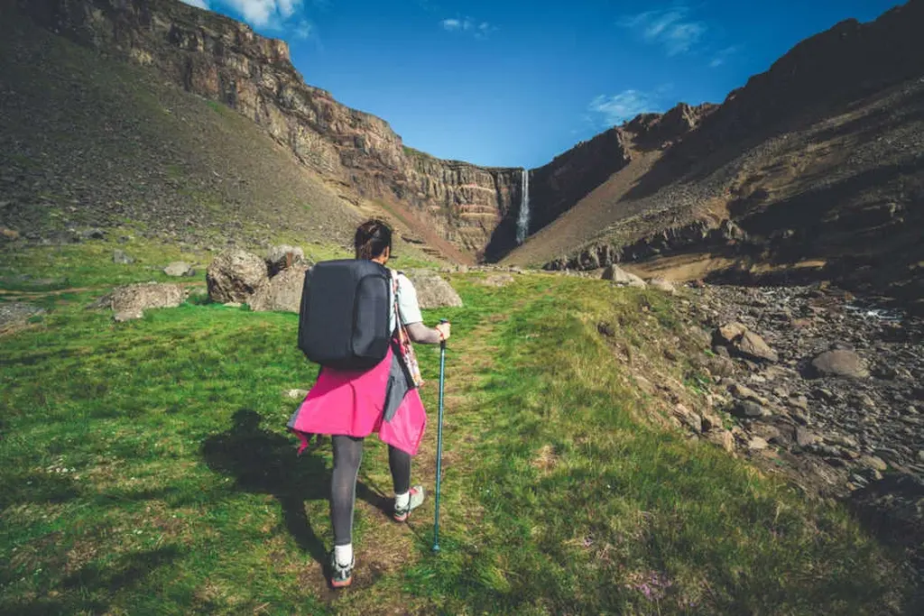 A hiker with a large backpack and trekking poles walking towards Hengifoss waterfall in Iceland. The path is surrounded by lush green grass, contrasting with the rocky terrain and towering canyon walls. The waterfall cascades down the cliff in the distance, under a bright blue sky with a few scattered clouds.