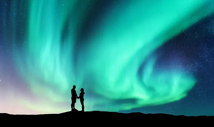 A silhouette of a couple standing hand in hand under the mesmerizing Northern Lights in Iceland. The sky is illuminated with vibrant green and blue auroras, creating a stunning natural light display. The couple stands on a hill, their figures dark against the bright and colorful sky, capturing a moment of awe and wonder. The stars are visible in the clear night sky, adding to the magical atmosphere of the scene.