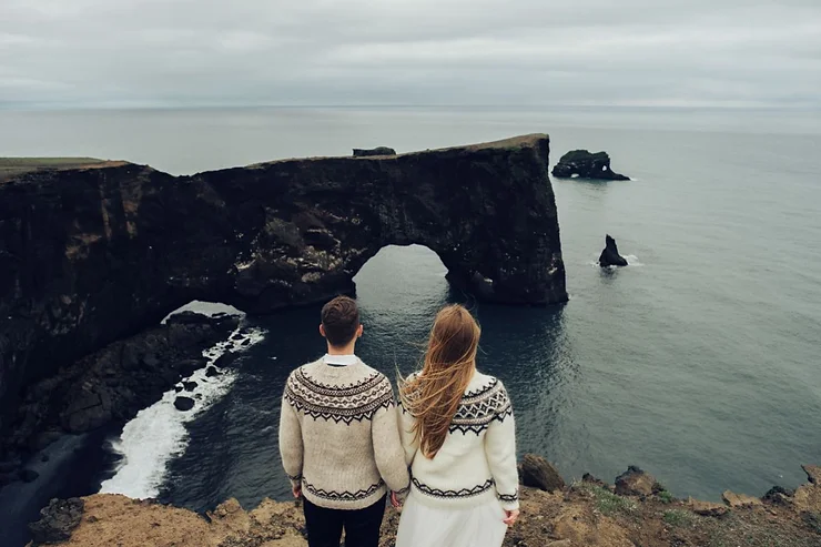 A couple in traditional Icelandic sweaters standing on a cliff overlooking the ocean in Iceland. They are gazing at a natural rock arch formation in the distance, surrounded by rugged coastal scenery. The overcast sky adds a dramatic touch to the serene and picturesque landscape. The couple's backs are to the camera, capturing a moment of shared tranquility and connection with nature.