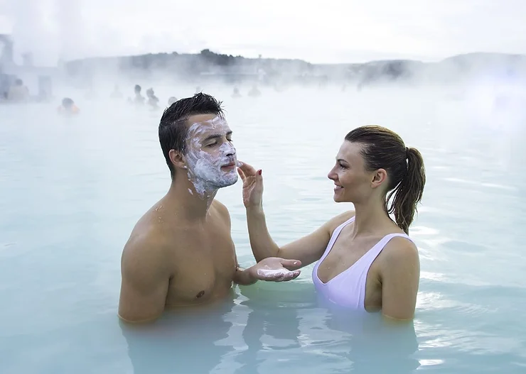 A couple enjoying a relaxing moment in a geothermal spa in Iceland. The woman is applying a face mask to the man while both are partially submerged in the milky blue waters. The background is filled with steam rising from the hot springs, creating a misty and serene atmosphere. Other spa-goers can be seen in the distance, enjoying the therapeutic waters. The couple is smiling and looking at each other, capturing a moment of intimacy and enjoyment.