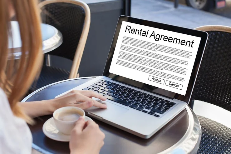 A woman sits at a round table with a cup of coffee while looking at a laptop screen displaying a car rental agreement. The agreement text is visible, with options to accept or cancel the agreement at the bottom of the screen. The background shows a blurred view of a cafe or public place, indicating a casual, relaxed setting.