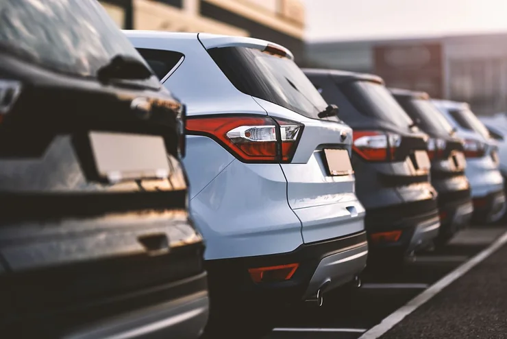 A line of parked rental cars, focusing on the rear ends of the vehicles. The cars are positioned closely together in a parking lot, with a variety of colors visible. The background shows a building and the soft light of either early morning or late afternoon, suggesting a busy rental location.