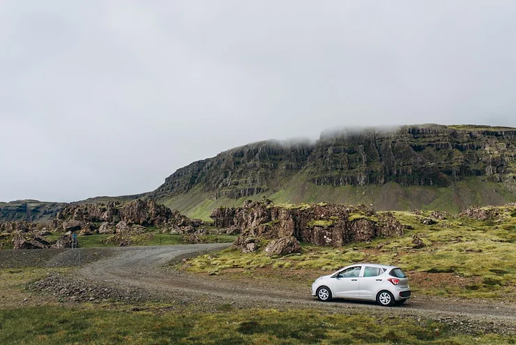 A white car is parked on a gravel road in Iceland, surrounded by a rocky and grassy landscape. The road winds through the terrain, leading towards steep, rugged cliffs in the background, partially obscured by mist. The scene is serene and remote, showcasing Iceland's natural beauty.