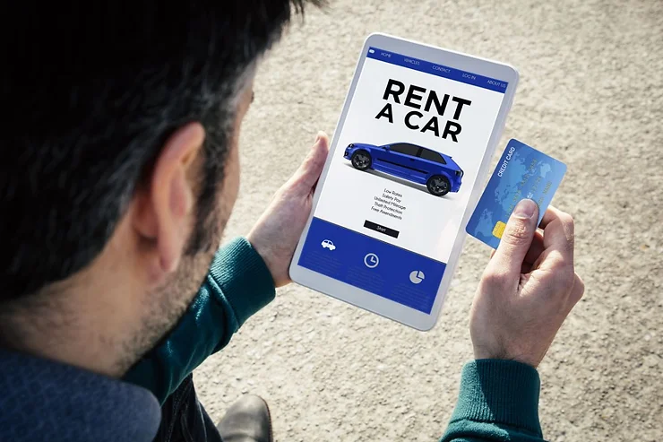 A man holds a tablet displaying a car rental app with the text "Rent a Car" and an image of a blue car. He is also holding a credit card in his other hand, preparing to complete the transaction. The tablet screen shows various rental options and buttons for selecting and confirming the booking. The background is blurred, focusing attention on the tablet and the man's hands.