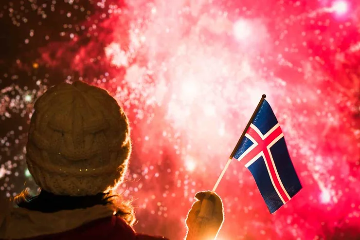 A person wearing a winter hat holds a small Icelandic flag against the backdrop of a vibrant red fireworks display. The warm glow of the fireworks illuminates the night sky, creating a festive and patriotic atmosphere as Reykjavik, Iceland, celebrates the New Year.