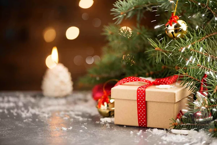 A small gift wrapped in brown paper and tied with a red polka-dot ribbon sits under a decorated Christmas tree. The tree is adorned with golden ornaments and fairy lights, creating a warm and cozy holiday ambiance. In the background, a candle softly glows, adding to the festive atmosphere.