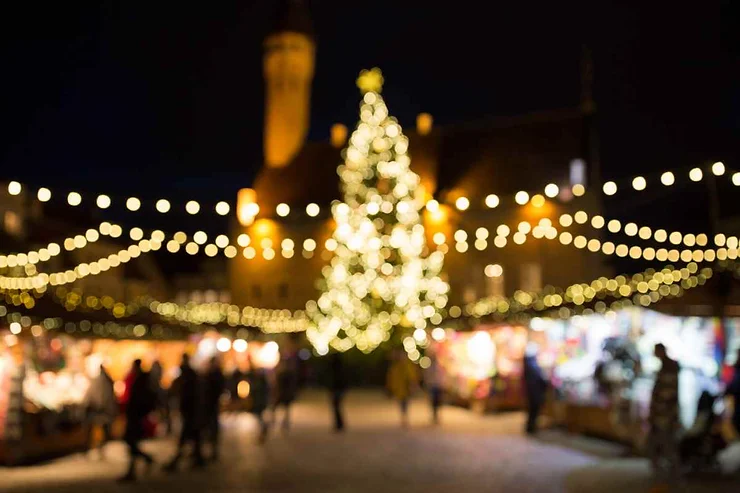 A beautifully lit Christmas tree stands at the center of a bustling Christmas market in Reykjavik, Iceland. The tree is adorned with sparkling lights and surrounded by festive string lights that illuminate the market stalls. People are seen strolling and enjoying the holiday atmosphere, with a historic building softly lit in the background, adding to the enchanting scene.