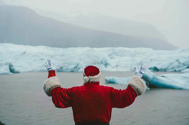 Santa Claus stands with his back to the camera, arms raised, in front of a stunning Icelandic glacier. The glacier's icy blue expanse contrasts with Santa's red suit and white fur trim. The misty, mountainous backdrop adds to the magical and festive atmosphere of the scene.