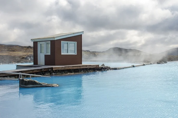 A tranquil scene at Lake Mývatn Natural Baths in Iceland, showcasing a small wooden cabin situated on the edge of the blue geothermal waters. Steam rises from the warm baths, blending with the overcast sky and creating a serene and inviting atmosphere. The surrounding landscape is rugged and serene, offering a peaceful retreat in nature.