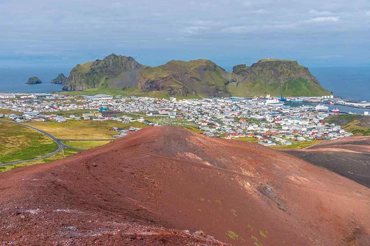 An aerial view of Heimaey in the Westman Islands, Iceland, showing the town spread out along the coastline. The foreground features a red volcanic landscape, while the background includes rugged green mountains and the blue ocean. The town's buildings and harbor are clearly visible, creating a striking contrast with the natural surroundings.