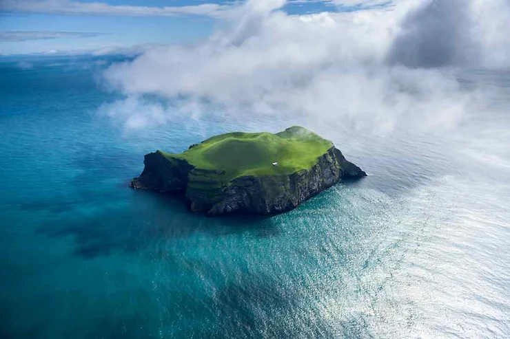 An aerial view of Heimaey Island in the Westman Islands, Iceland. The small island is covered in lush green grass and is surrounded by the deep blue waters of the ocean. Clouds partially obscure parts of the island, adding to the sense of isolation and natural beauty.