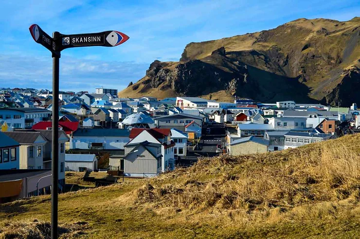 A picturesque view of Heimaey in the Westman Islands, Iceland, featuring a quaint town with colorful rooftops nestled against a rugged hillside. A signpost in the foreground points towards Skansinn, adding a touch of local charm. The clear blue sky and the natural landscape provide a stunning backdrop to the serene and vibrant community.