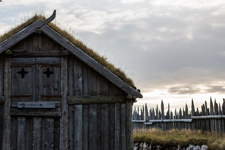 Historic Icelandic wooden building with a turf roof, featuring a rustic gate and a fence with pointed stakes against a cloudy sky.